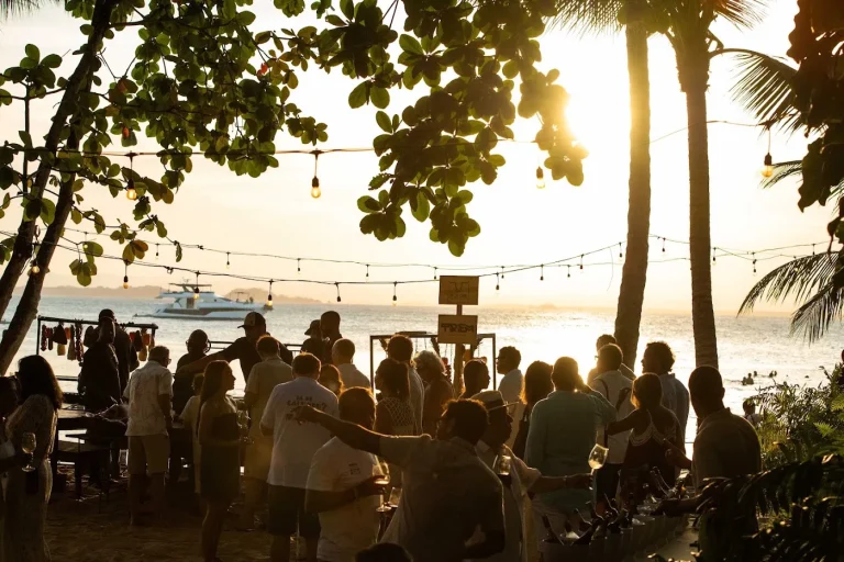 Convidados reunidos sob as árvores e o varal de luzes, virados para o mar no pôr do sol