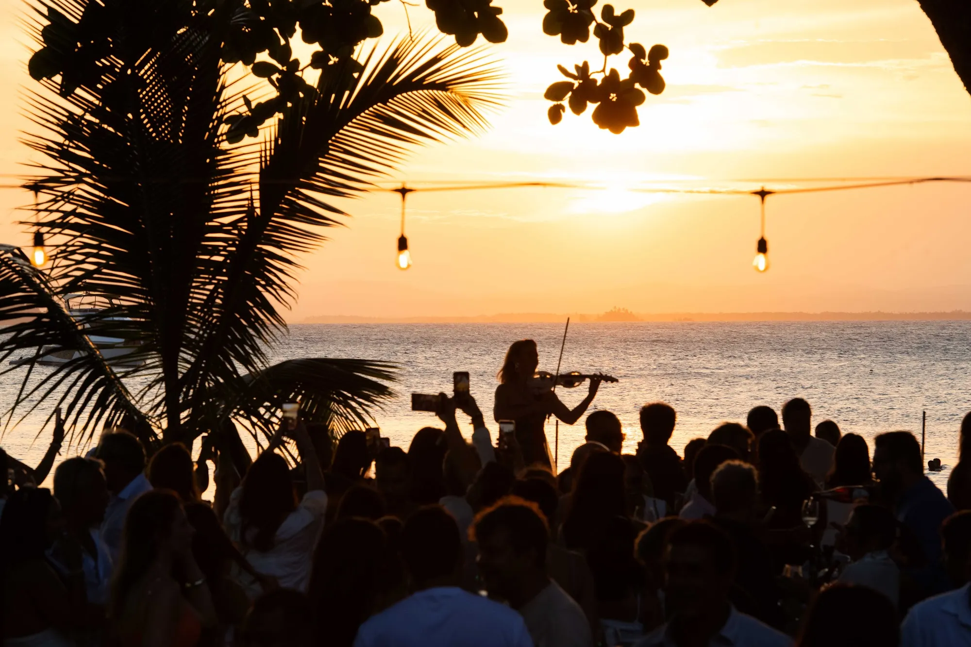 Pôr do sol sobre a Baía de Camamu durante o Wine Festival Obar 2026: violinista e convidados em silhueta diante do mar dourado
